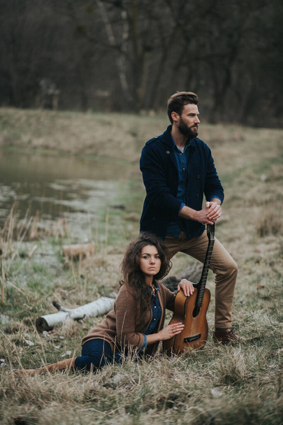 δύο καυκάσιους εραστές μουσικούς με κιθάρα να στέκονται δίπλα στη λίμνη. Νεαρό ζευγάρι αγκαλιάζεται την ημέρα του φθινοπώρου έξω. Ένας γενειοφόρος άντρας και μια σγουρή γυναίκα ερωτευμένη. Του Αγίου Βαλεντίνου. Έννοια της αγάπης και της οικογένειας - Φωτογραφία, εικόνα