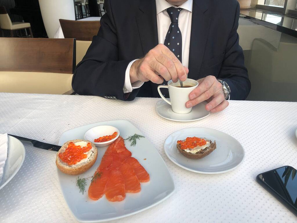 Takım elbiseli, kravatlı ve beyaz gömlekli bir adam kahvaltı, kahve molası, bir fincan kahve, somonlu kruvasan ve yeşil salatalı sandviç ve havyarlı sandviç yiyor. - Fotoğraf, Görsel