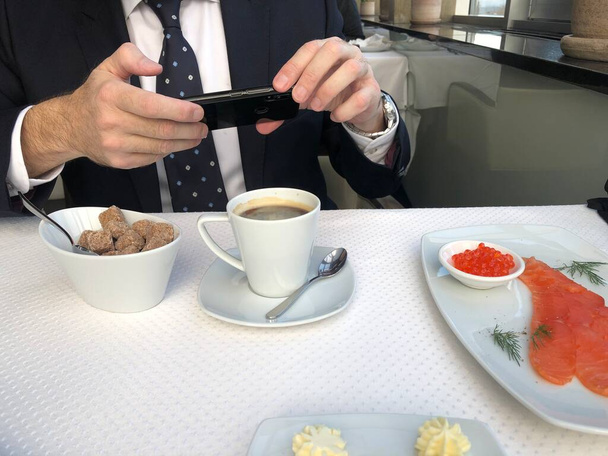 Muž v obleku a kravatě používající chytrý telefon u jídelního stolu. Užívá si sociálních médií a snídaně. - Fotografie, Obrázek