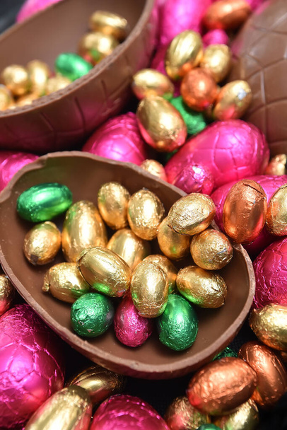 Pile or group of multi colored and different sizes of colourful foil wrapped chocolate easter eggs in pink, red, gold and lime green with two halves of a large brown milk chocolate egg in the middle and mini eggs inside. - Photo, Image