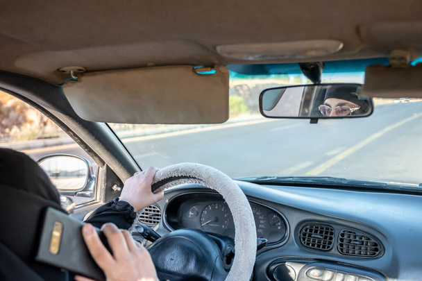 Mujer árabe musulmana conduciendo un coche mientras usa su teléfono inteligente - Foto, Imagen