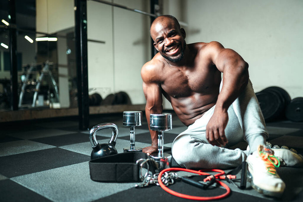 A man with a naked torso in gray pants sits on the floor next to dumbbells and a kettlebell. Strength training using sports equipment - 写真・画像