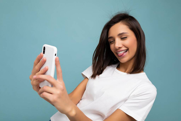 Foto van sexy mooi glimlachen gelukkig jong brunette vrouw dragen casual wit t-shirt geïsoleerd over muur blauwe achtergrond houden en met behulp van mobiele telefoon het nemen van selfie kijken naar gadjet scherm en - Foto, afbeelding