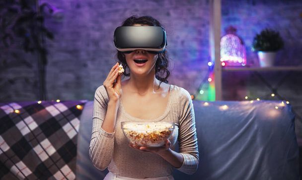 Νεαρό κορίτσι που παίρνει εμπειρία VR ακουστικά χρησιμοποιεί γυαλιά επαυξημένης πραγματικότητας είναι στην εικονική πραγματικότητα. Νεαρή γυναίκα φορούν VR ακουστικά και να παρακολουθήσουν ταινία με ποπ κορν στο σπίτι διαμονή - Φωτογραφία, εικόνα