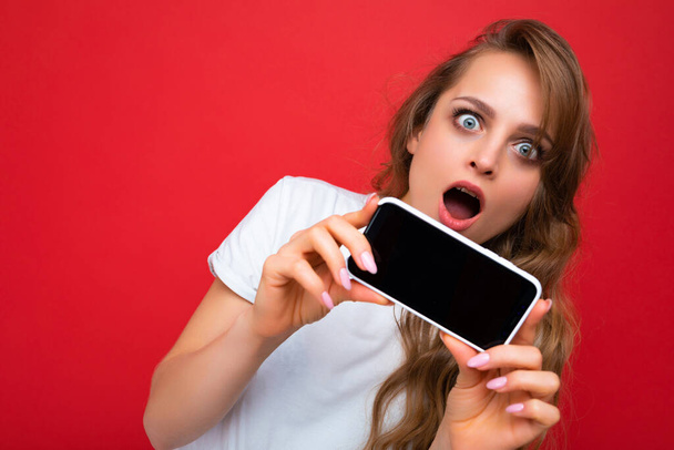 Удивлённый шокированный красивая улыбающаяся молодая блондинка, симпатичная в белой футболке, стоящей изолированно на красном фоне с копией пространства с телефоном в руке со смартфоном с пустым экраном - Фото, изображение