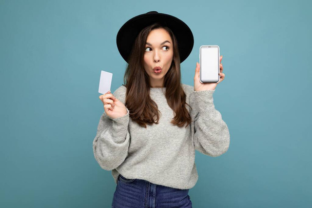 Photo of attractive amazed young brunette woman wearing black hat and grey sweater isolated over blue background holding credit card and mobile phone with empty display for mockup looking to the side - Photo, Image