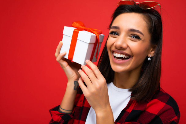 Girato di attraente positivo sorridente giovane donna bruna isolata su sfondo colorato parete indossare ogni giorno vestito alla moda in possesso di scatola regalo e guardando la fotocamera - Foto, immagini