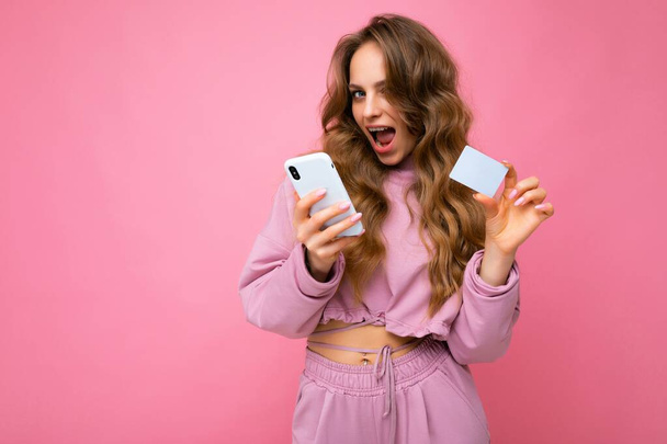 Mooie positieve jonge blonde krullende vrouw dragen roze kleren geïsoleerd over roze achtergrond met behulp van mobiele telefoon betalen online winkelen via credit card kijken naar camera - Foto, afbeelding