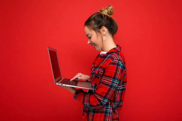 profilo laterale foto di bella giovane che tiene netbook guardando schermo digitando sulla tastiera indossando camicia rossa isolato su sfondo muro rosso - Foto, immagini