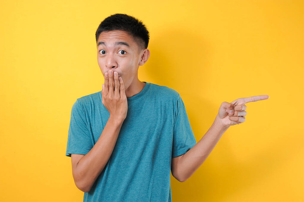 Hombre adolescente poniendo el dedo en la boca un signo de gesto de silencio apuntando al espacio de copia para mostrar algo, aislado sobre fondo amarillo - Foto, imagen
