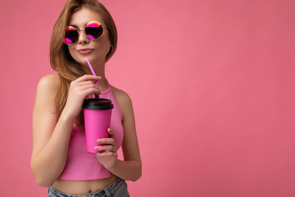 Очаровательная сексуальная молодая счастливая блондинка, одетая в стильную одежду, изолированную над красочной стеной на заднем плане, держащая бумажный стаканчик для макета, пьющая молочный коктейль, глядя в сторону - Фото, изображение