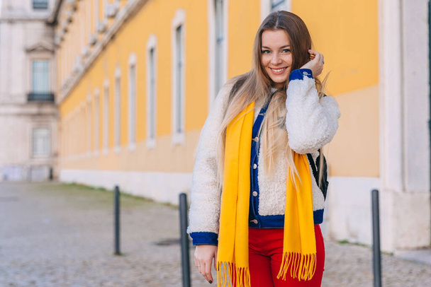 Девушка с длинными волосами смотрит в камеру и улыбается на заднем плане здания с желтым фасадом. Закрывай. стильная одежда. - Фото, изображение