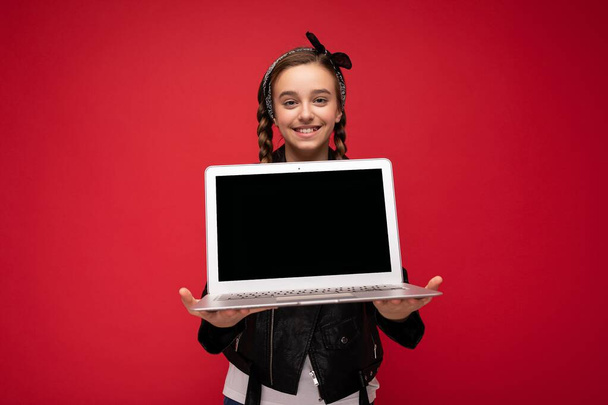 Photo of beautiful happy smiling girl with brunet pigtails holding computer laptop wearing black jacket and bandana isolated over red wall background looking at camera - Foto, immagini