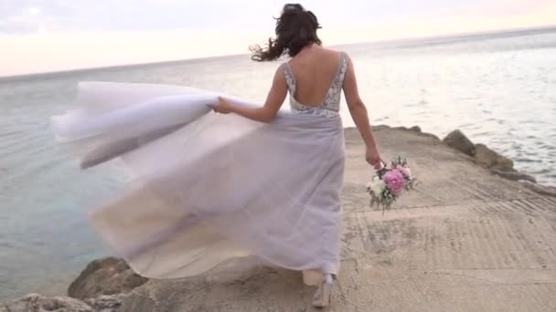 la novia con un ramo de bodas camina por el muelle, sosteniendo su falda con su mano - Imágenes, Vídeo