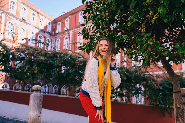 dziewczyna w stylowych ubraniach pozowanie w pobliżu drzewa na tle budynku z czerwoną fasadą. Lizbona, Portugalia. - Zdjęcie, obraz