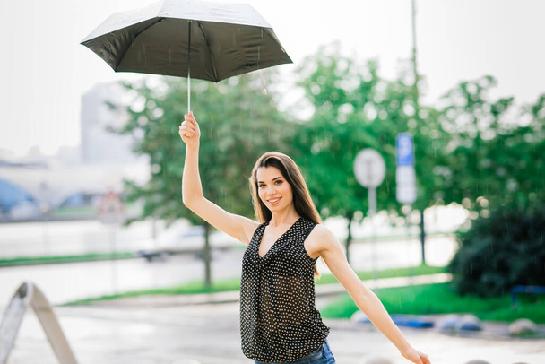 Portret van een gelukkige vrouw in jeans onder een paraplu lachend op straat in regenachtige dagen - Foto, afbeelding