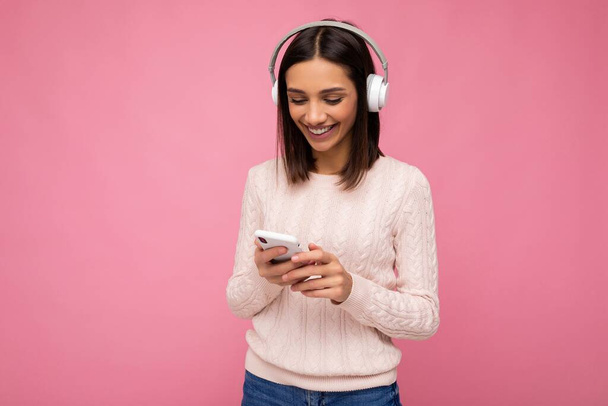 Photo shot of beautiful joyful smiling young female person wearing stylish casual outfit isolated over colorful background wall wearing white bluetooth wireless earphones and listening to music and - Foto, imagen