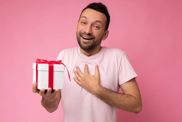 Fototermin von hübsch positivem Lächeln brünette unrasierte junge Mann isoliert über rosa Hintergrund Wand trägt rosa T-Shirt hält weiße Geschenkbox mit roter Schleife und Blick in die Kamera - Foto, Bild