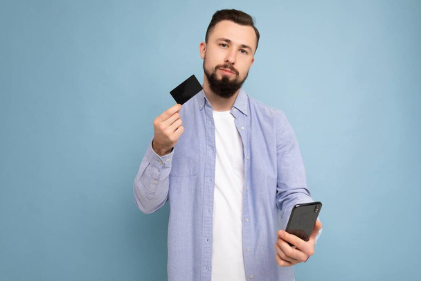 Fénykép a figyelmes jóképű vonzó barna borotválatlan fiatalember visel alkalmi kék ing és fehér póló elszigetelt kék háttér fal gazdaság hitelkártya és a mobiltelefon-készítés - Fotó, kép