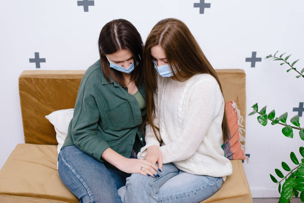 Młode kobiety w maskach medycznych siedzą na kanapie i trzymają się za ręce - Zdjęcie, obraz