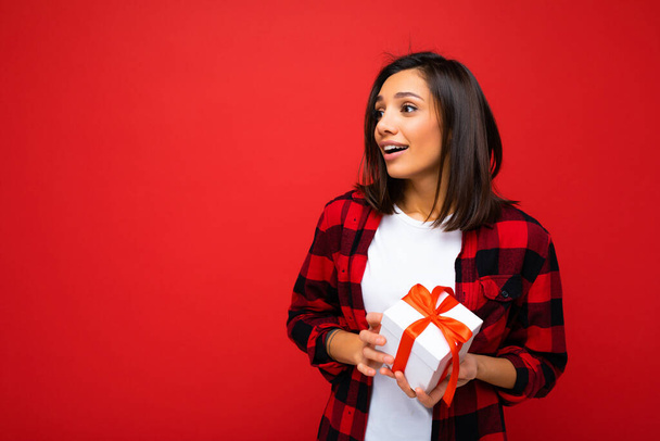 Photo shot of pretty positive young brunette woman isolated over red background wall wearing white casual t-shirt and red and black shirt holding white gift box with red ribbon and looking to the side - Photo, Image