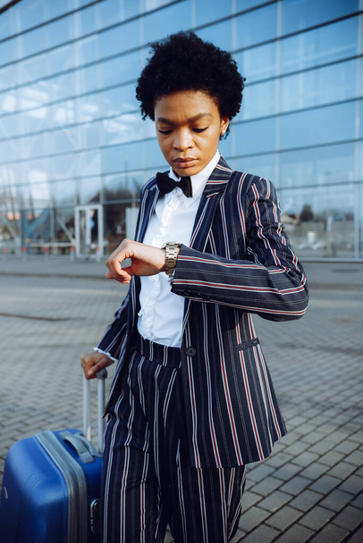 4k. Reisen, Digital. Charmante Afroamerikanerin im modern gestreiften Anzug steht an der Glaswand neben ihrer Reisetasche. Unternehmerin oder Studentin. Blicke auf Uhren. - Foto, Bild