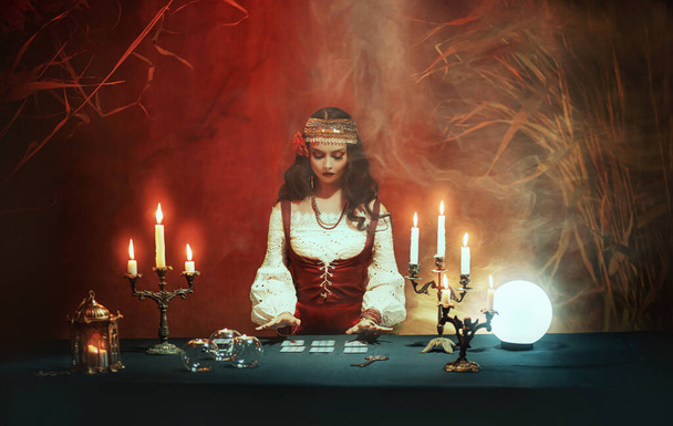 Fantazie dívka v obrazu cikánské čarodějnice sedí u stolu v tmavě gotické místnosti. Kostým Art Red. Kouzelná žena věštkyně čte budoucnost na tarotových kartách, křišťálová koule. Rituální hořící svíčky, seance, kouř - Fotografie, Obrázek