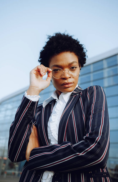 4k. Charming African American woman in modern striped suit stands on background of glass wall in elegant pose, touches by arm her glasses standing before a modern glass building and looks on camera - Fotó, kép