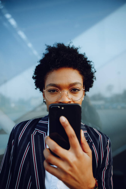 4k. Charming African American woman in modern striped suit. Holding her phone looks in camera. Businesswoman or student. Close up. - Photo, Image