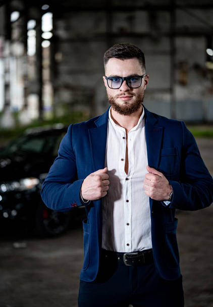 Handsome man poses in blue suit and white shirt on old storehouse background. Modern car behind model boy. - Zdjęcie, obraz