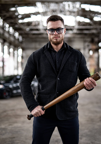 Cool brutal guy with baseball bat in hands. Total black outfit on abandoned warehouse background. Brutal man concept. - Foto, Bild