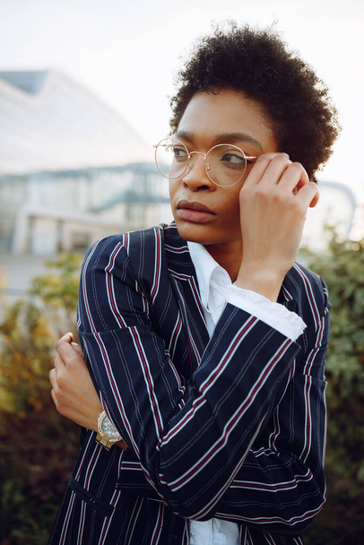 4k. Travel, Digital. Charming African American woman in elegant striped suit, looks on left side, touches her glasses. Stands on background the bushes near airport. Businesswoman or model. Garden - Φωτογραφία, εικόνα