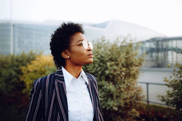 4k. Travel, Digital. A charming African American woman in an elegant striped suit, looks at the sky, and waiting for her flight. She stands on background of bushes near the airport. Businesswoman or - Zdjęcie, obraz