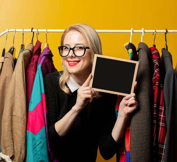 Style woman in black formal dress hold board next to clothes on rail hanger on yellow background - 写真・画像