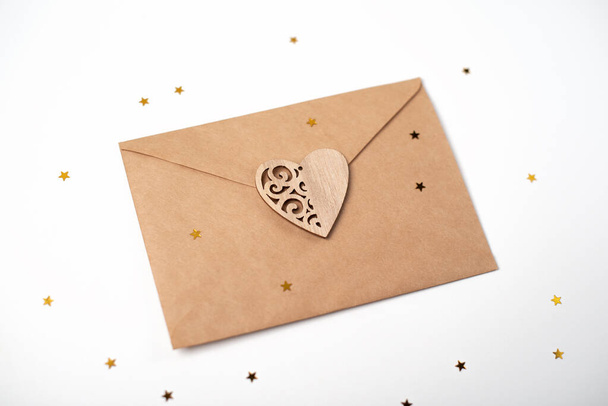 Craft envelope with the wooden heart and little golden stars on the light (white) background. Flat lay, top view. Romantic love letter for Valentine's day concept. - Foto, immagini