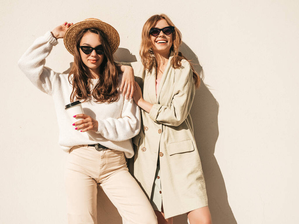Two young beautiful smiling hipster female in trendy white sweater and coat. Sexy carefree women posing near wall in the street in hat and sunglasses. Positive models having fun and drinking coffee  - Фото, зображення