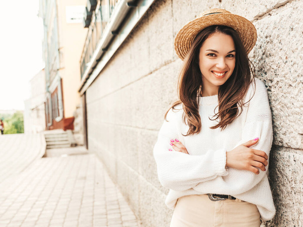 Portrait of beautiful smiling cute model. Female dressed in warm hipster white sweater and hat. Posing near wall in the street. Funny and positive woman hugging herself  - Zdjęcie, obraz