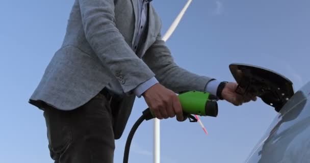 Muž připojí nabíječku ke svému elektrickému autu na pozadí rotujících lopatek větrného mlýna, zapojí ho a ustoupí stranou.. - Záběry, video