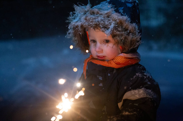 Маленькая девочка в зимней одежде, выходящая на улицу и держащая искру в руке. Темно и снежно, девушка счастливо улыбается. Волшебное зимнее настроение - Фото, изображение