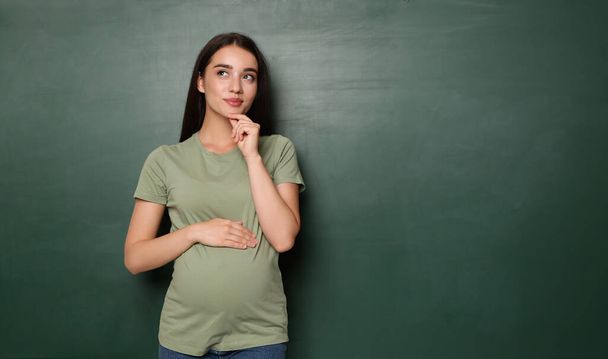 Pregnant woman near empty green chalkboard, space for text. Choosing name for baby - Zdjęcie, obraz