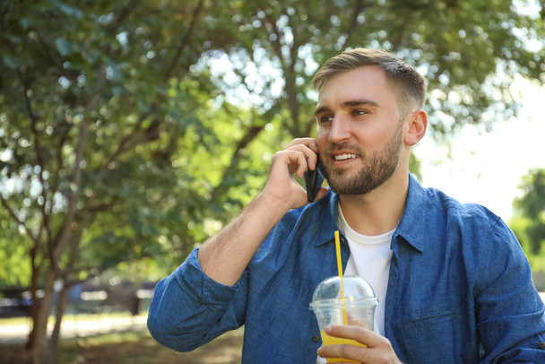 Young man with refreshing drink talking on smartphone in park - Fotografie, Obrázek
