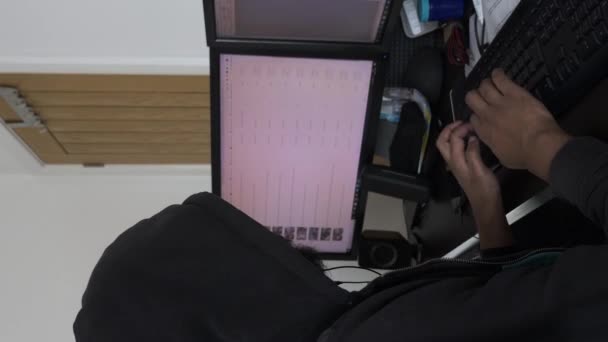 Hombre con capucha escribiendo delante de los monitores duales en el Ministerio del Interior. Vídeo vertical, bloqueado - Metraje, vídeo