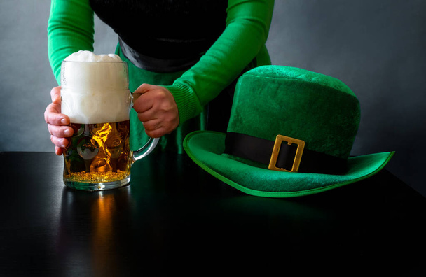 Main de femme tenant un verre de bière près du chapeau de Saint-Patrick d'un lutin sur le fond noir - Photo, image