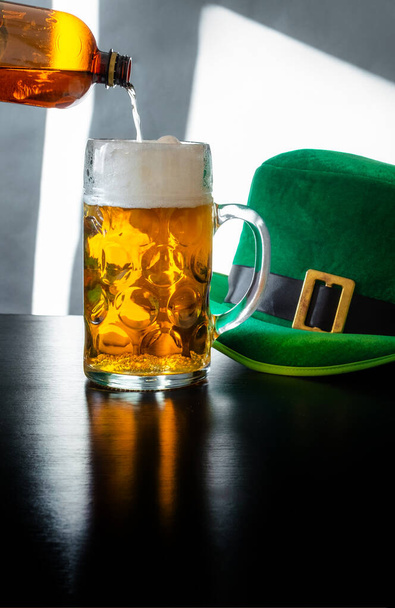 Woman's hand holding glass of beer near St. Patrick's day hat of a leprechaun on the black background - Foto, afbeelding