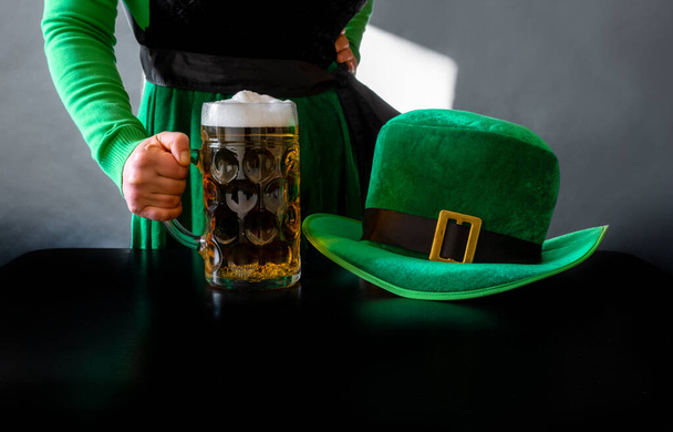 Die Hand einer Frau hält ein Glas Bier in der Nähe von St. Patrick 's Day Hut eines Kobolds auf schwarzem Hintergrund - Foto, Bild