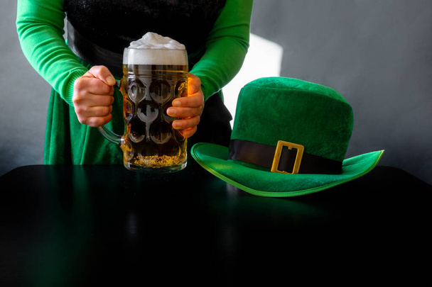 Woman's hand holding glass of beer near St. Patrick's day hat of a leprechaun on the black background - Foto, imagen