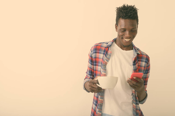 Studio strzał młody szczęśliwy czarny Afrykanin uśmiecha się i trzyma filiżankę kawy podczas korzystania z telefonu komórkowego na białym tle - Zdjęcie, obraz
