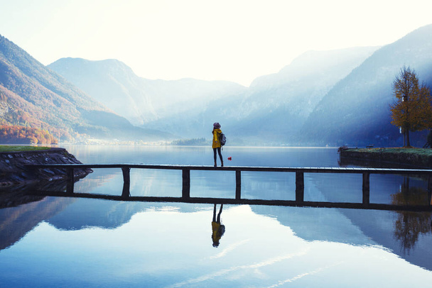 girl tourist in a hat and with a backpack stands on a wooden bridge on a mountain lake in the early morning. beautiful landscape and reflectio - Foto, Imagen