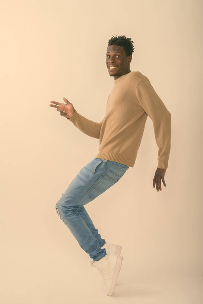Plan complet du corps de jeune homme africain noir heureux souriant tout en posant et en tiptoeing sur fond blanc - Photo, image