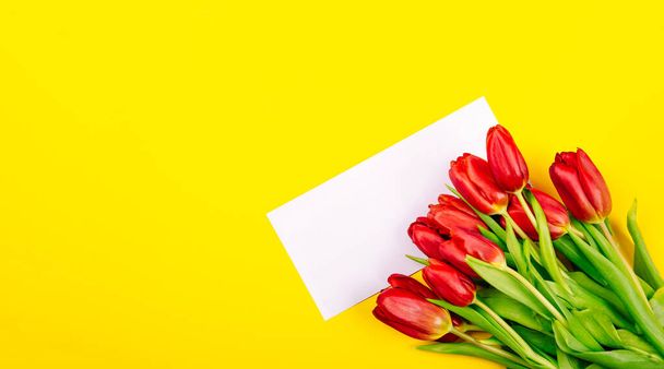 Witte cadeaubon envelop of visitekaartje op gele achtergrond met een boeket rode tulpen en kopieer ruimte, tekst plaats. Een kerstkaart. Gelukkige Valentijnsdag of Vrouwendag of Moederdag. - Foto, afbeelding